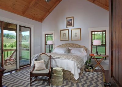 This vintage rustic bedroom features white walls and exposed beams, creating an elegant rustic-like ambiance. The bed is adorned with light colored sheets one with flower designs. A large light colored patterned rug adds distinction on the dark hardwood floor. Natural light beams through glass doors and and windows, enhancing the inviting atomosphere. A bedside table with a lamp and books contributes to the room's charm, while vintage elements, like an old drawing framed by golden trim on the wall and a vintage looking chair in the front of the bed, complete the vintage-inspired look.