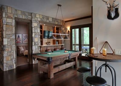 Rustic game room with a wooden pool table, stone walls, and a hanging light fixture over the table; bar seating and a doorway to a deck outside.