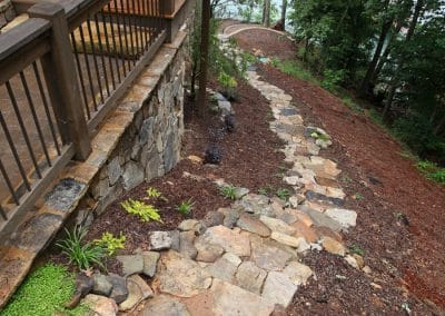 Stone stepping-stone path along a sloped yard beside a wooden deck, winding toward a lakeside with trees in the background.