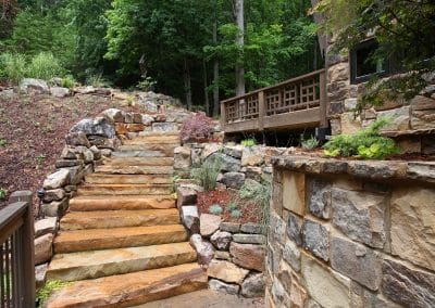 Stone steps and layered rock walls ascend through a landscaped yard beside a stone house and wood-deck veranda in a wooded setting.