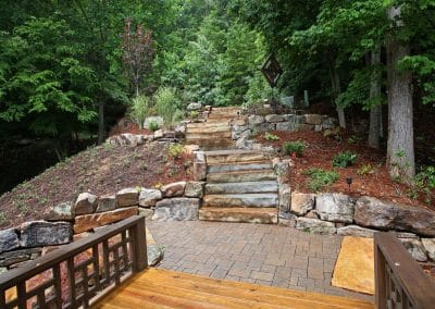 Stone staircase and layered rock walls rise through a landscaped backyard with trees and mulch, seen from a wooden deck.