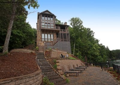Lakeside multi-story stone-and-wood house with decks and stairways, overlooking a stone-patio terrace with chairs by the water.