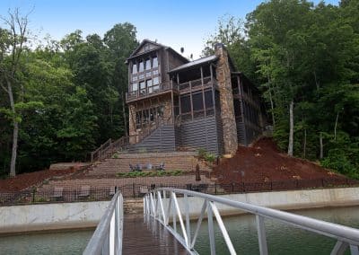 Three-story wood-and-stone lodge perched on a hillside above a lake, with a metal bridge in the foreground leading to it.