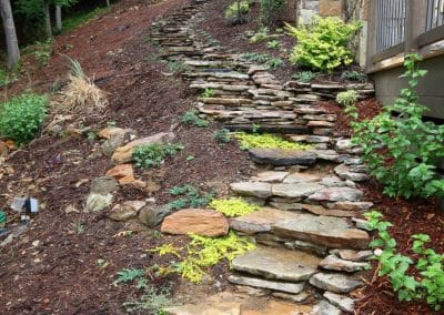 Stone stepping-stone path winding up a sloped garden beside a house with a stone retaining wall and wooden deck.
