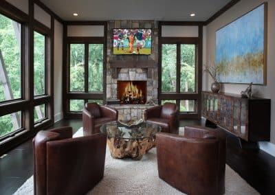 Cozy modern living room with a stone fireplace and a wall-mounted TV, surrounded by four brown leather chairs around a glass-top coffee table on a white rug.