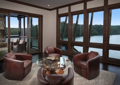Cozy living room with brown leather chairs around a glass-top table, looking out to a lake through large wooden-framed windows.