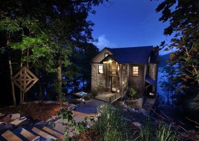 Stone-and-wood cabin lit at dusk, perched on a rocky hillside with trees and a lake in the background.