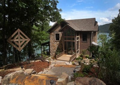 Lakeside stone-and-wood house with large glass entrance and wooden deck, overlooking a blue lake with surrounding trees and rocks ahead.