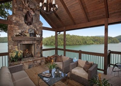 Lakeside covered porch with a stone fireplace, mounted deer head, and wicker seating overlooking the lake.