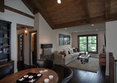 Living room with exposed wooden beams, a large window, and a beige sectional sofa; a round dining table shows a tic-tac-toe game on wooden pieces in the foreground.