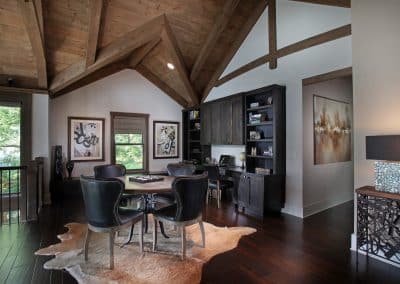 Rustic-modern home office with exposed wooden beams, a dark wood built-in cabinet and shelves, and a round table surrounded by four black leather chairs on a cowhide rug.