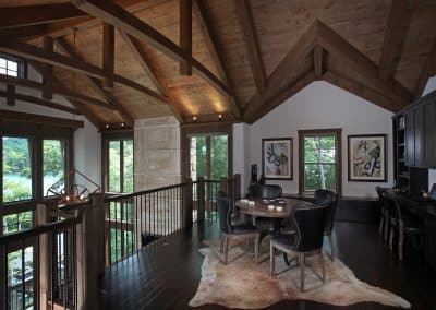 Rustic loft living area with exposed timber beams, a round wooden table and five black leather chairs on a cowhide rug, overlooking a lake through large windows.