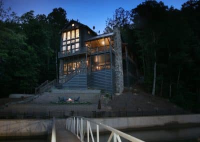 Lakeside house at dusk with warm interior lights, large windows, stone chimney, and multiple decks overlooking a terraced yard and water.