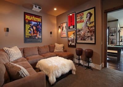 Cozy basement living room with a tan sectional sofa, white fluffy ottoman, vintage movie posters, and a ceiling projector.